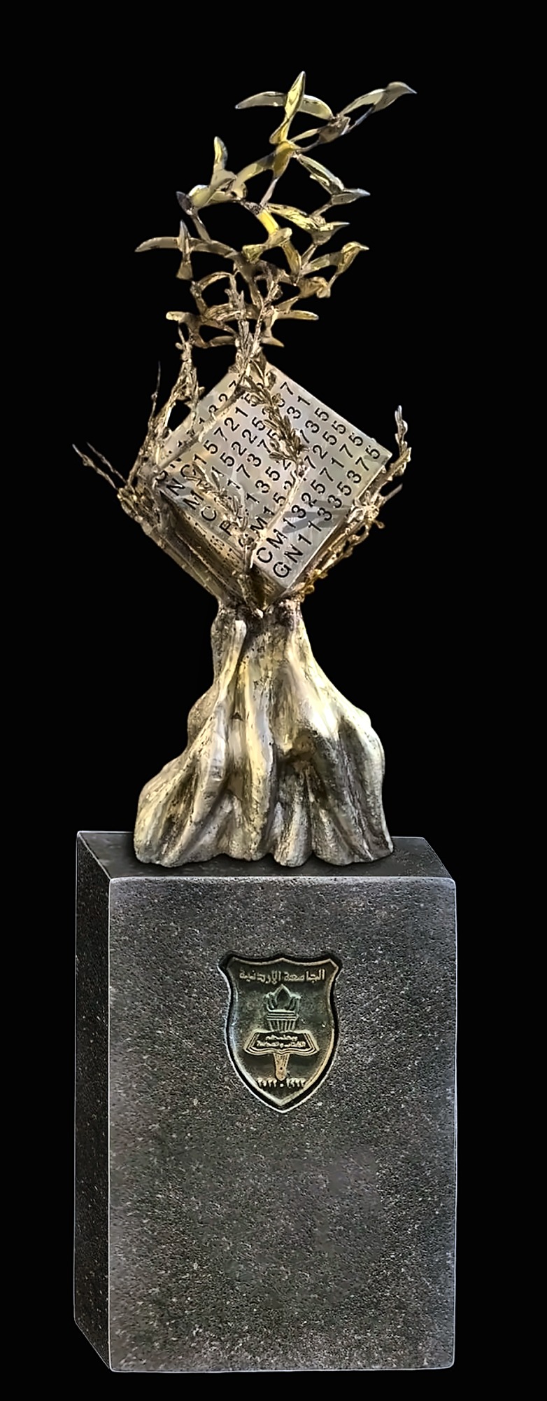 The same bronze olive tree and numbered cube, photographed against black on a dark stone plinth bearing the crest of the University of Jordan.