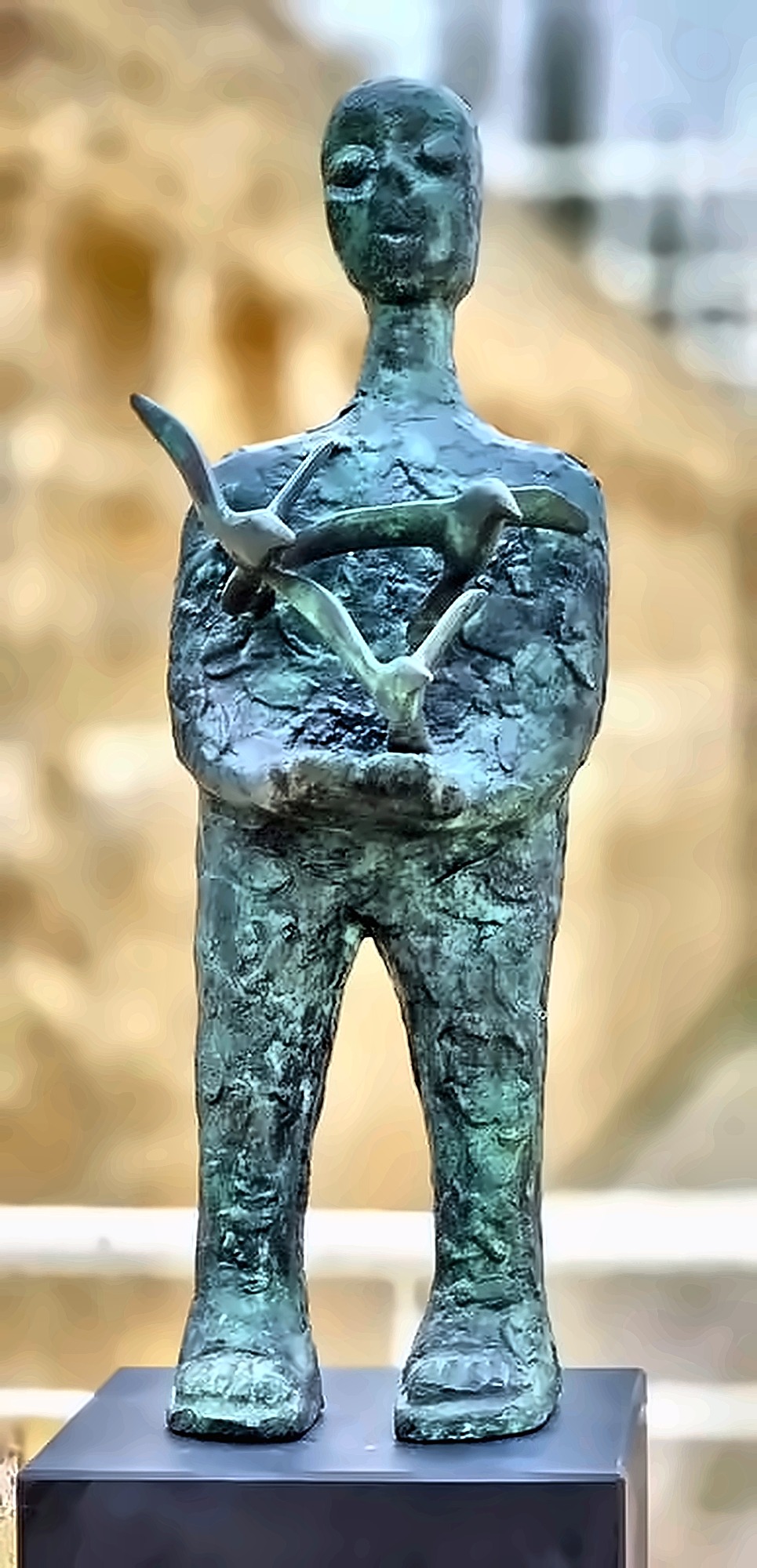 A green-patinated bronze figure standing with both arms curved at the waist, three birds rising out of its cupped hands.