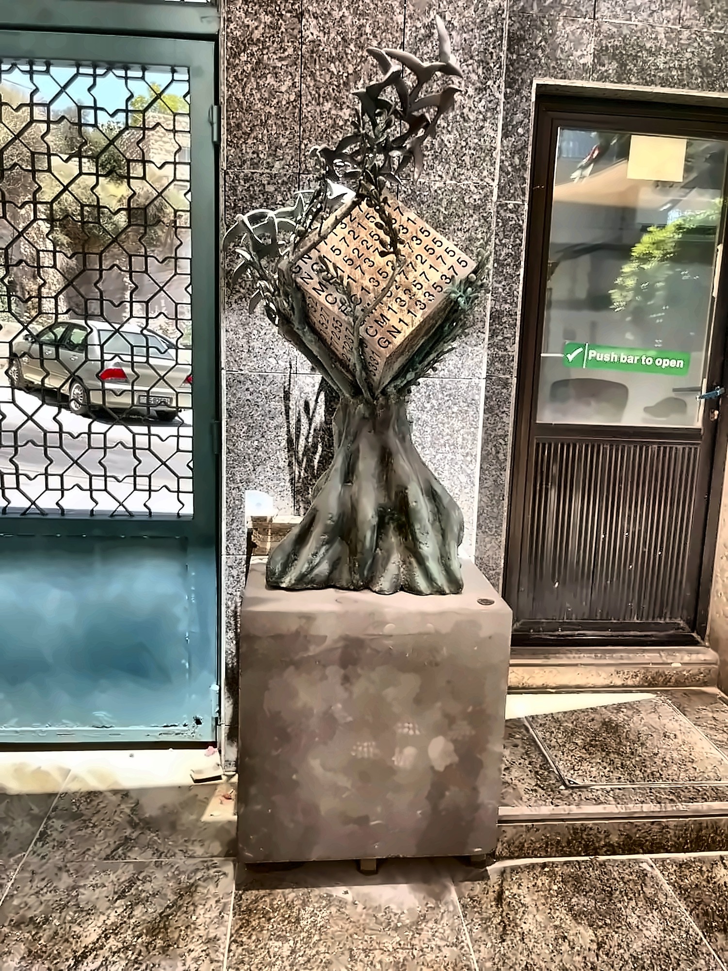 A bronze olive tree on a stone plinth, holding a cube incised with serial numbers, birds rising from the branches, standing inside a doorway of the Central Bank of Jordan.