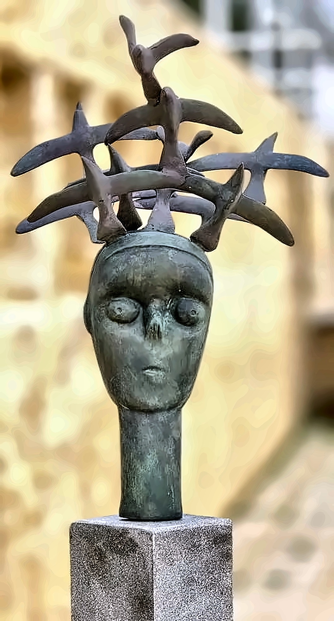 A bronze head on a stone plinth wearing a crown of birds set in a flat ring, wings overlapping.