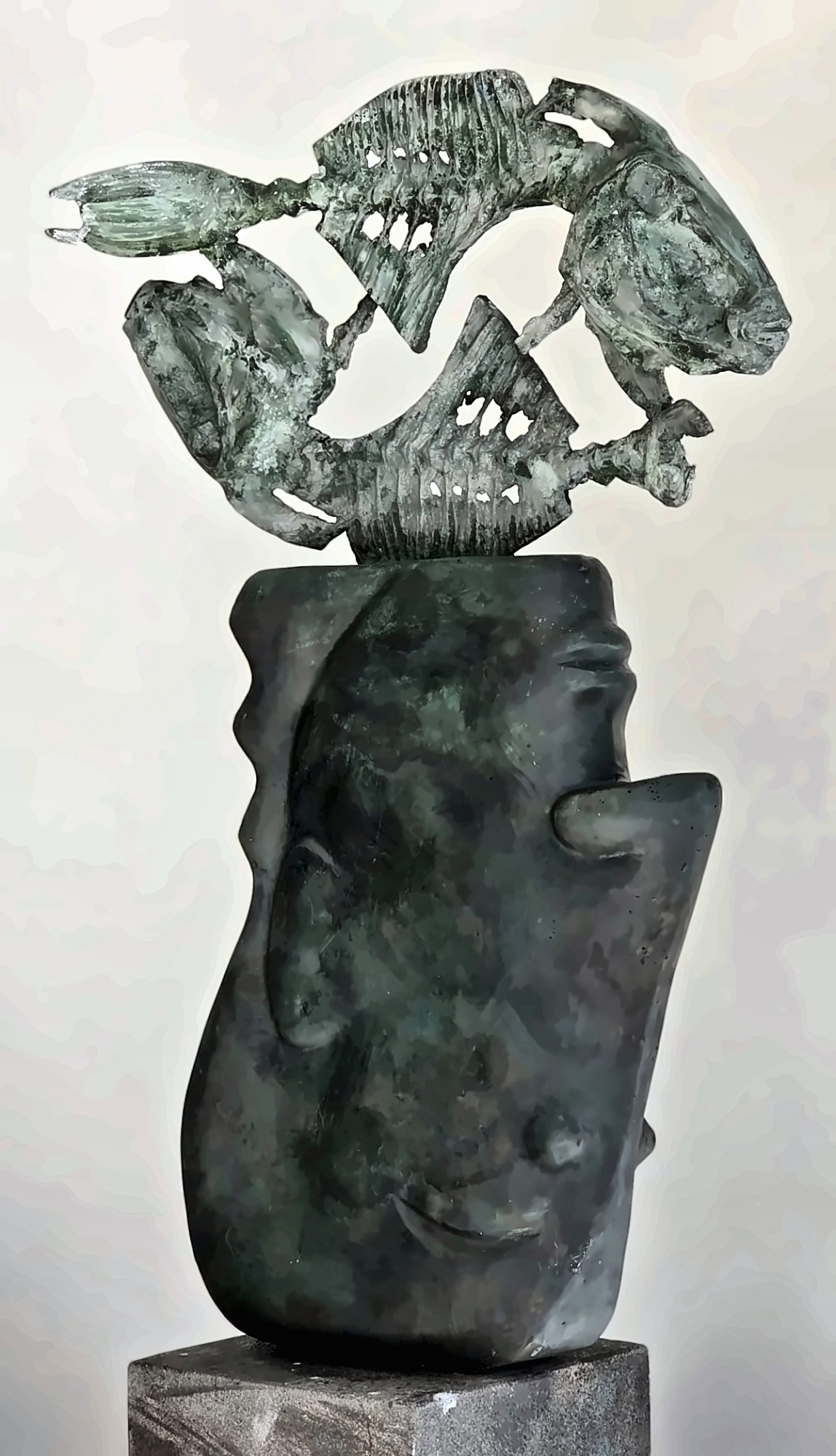 A green-patinated bronze head with pierced fish forms rising above it on a stone plinth.