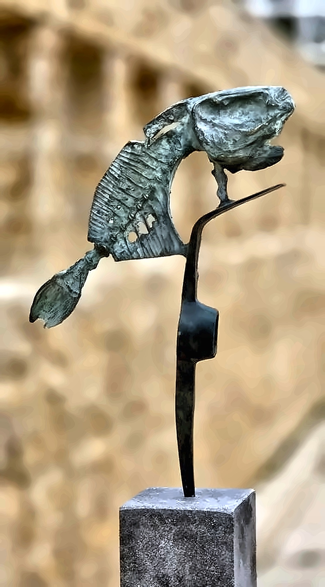 A pierced bronze fish balanced on a thin black steel stalk on a stone plinth.