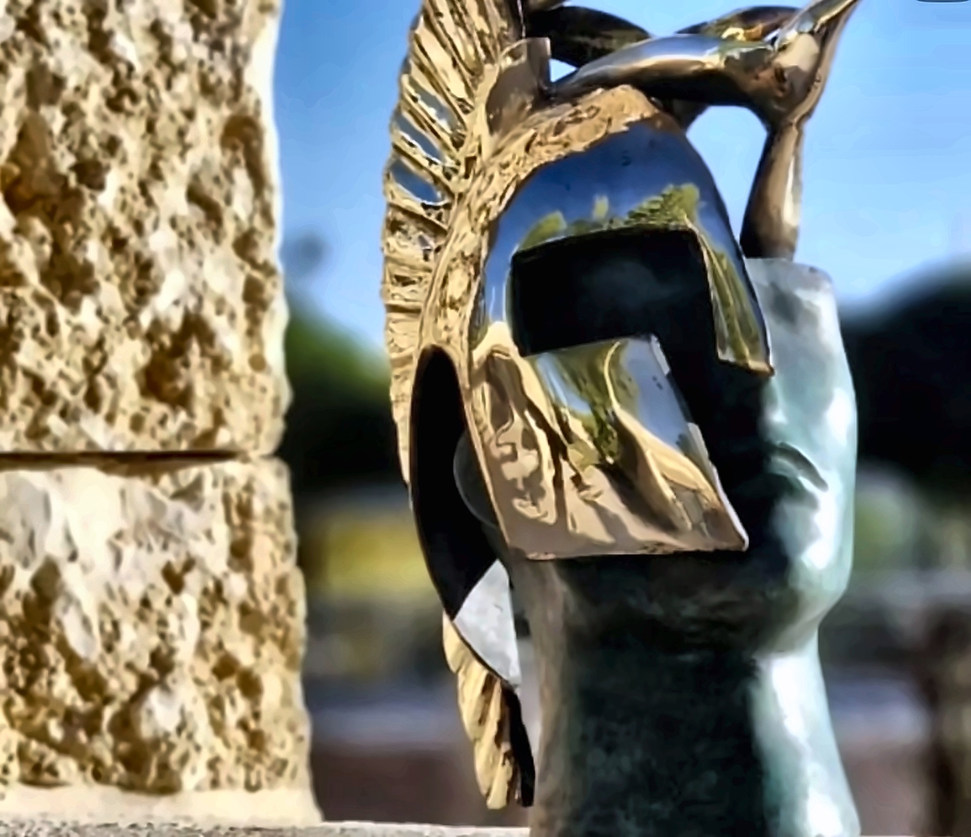 A stone form fitted with a bronze helmet and a bird, against a stone wall.