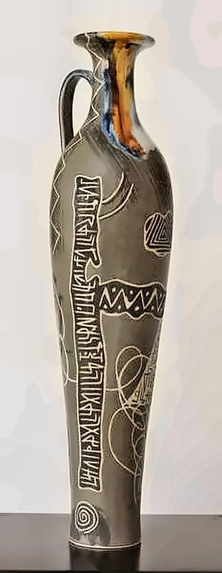 A tall grey jug with a handle, incised with vertical lines of script, colour running at the neck.
