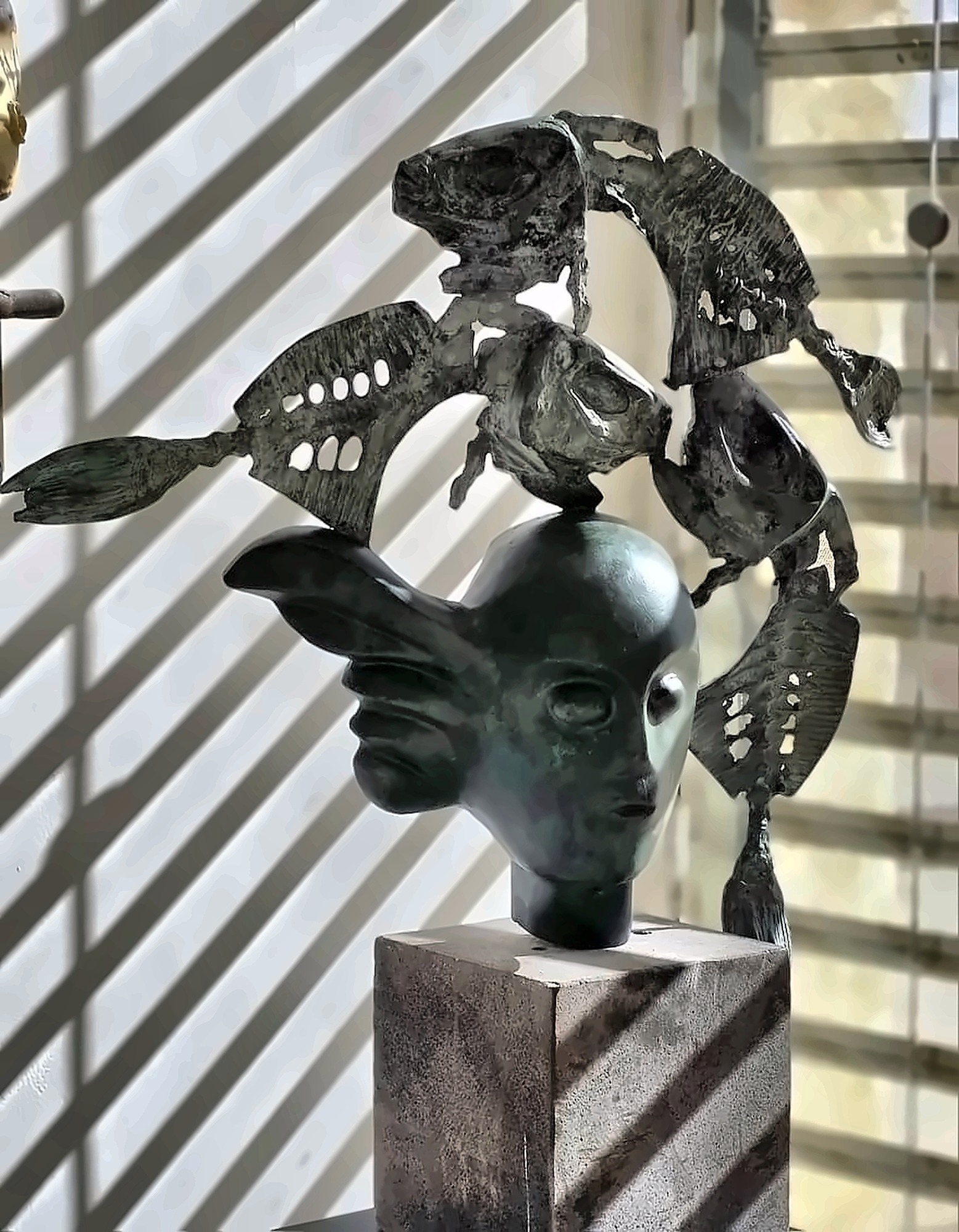 A second bronze head, this one winged, with pierced fish above it, in light thrown through window blinds.